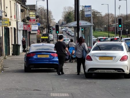 Two premium cars parked on the pavement in front of shops on Tong Street whilst pedestrians attempt to navigate around