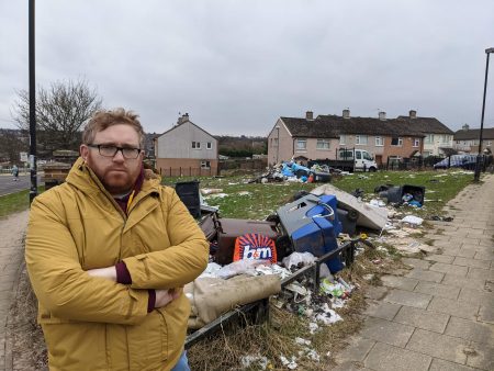 Matt Edwards standing next to a large amount of fly tipped rubbish outside houses on Melcombe Walk in Holmewood, Bradford.