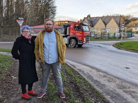 Celia Hickson and Matt Edwards standing outside Woodlands Primary School (an small Victorian style school building) with a large red lorry on the road behind them. The grass verge has been damaged by vehicles parking on it.