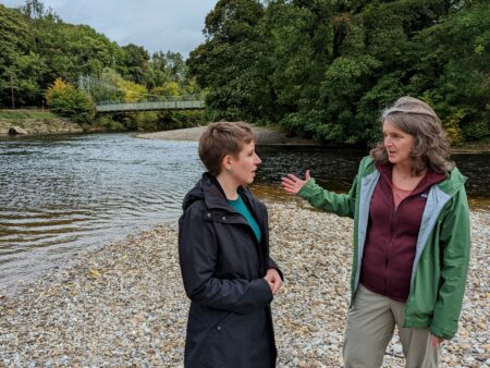 Carla Denyer and Ros Brown talking to each other on the banks of the River Whatfe. There are a lot of trees on both sides of the river and a small suspension bridge in the background.