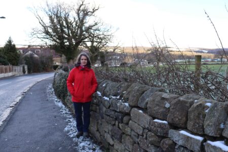 Ros Brown standing on a pavement next to a dry stone fence with a field in the background. To the left there is a road.