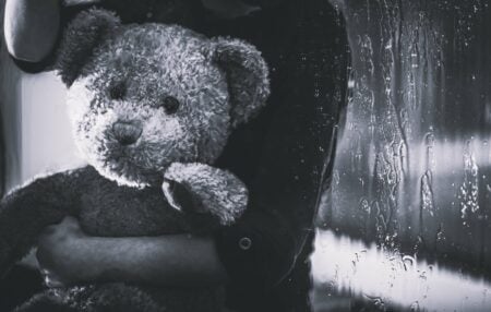 Black and white photograph of a children's teddy being held by a child. In the background is a window with rain.