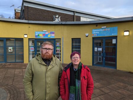 Councillors Matt Edwards and Celia HIckson stood outside the front of Holmewood Library. A curved building with yellow painted walls