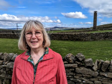 Councillor Janet Russell wearing a pink coat standing with a view of countryside between Silsden and Addingham in the vackground. There is a blue sky with a few clouds.