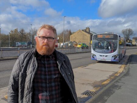 Councillor Matt Edwards standing at a bus stop with a single decker grey First Bus in the background approaching the bus stop.