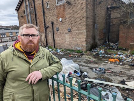 Councillor Matt Edwards standing outside the Holmewood Social Club, a 50s era brick building/ There is a large amount of waste dumped outside.