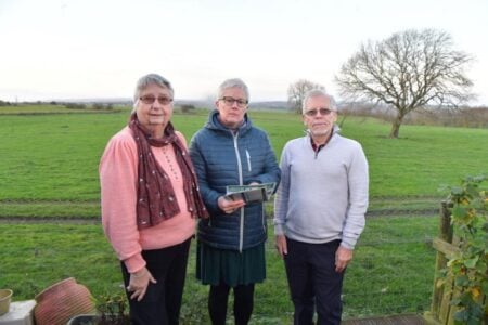 Councillor Celia Hickson with residents Iain and Diane McInnes with the development site behind. It is an area of green field with trees.